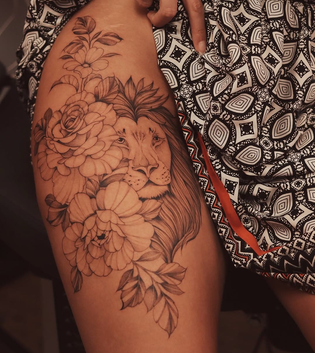 This tattoo for Emma features a majestic lion’s face surrounded by detailed roses and peonies. The design, symbolizing strength and elegance, is beautifully shaded and positioned along her thigh, creating a bold yet graceful statement that complements her leg’s contours.