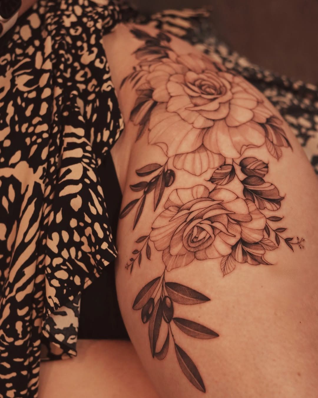 This is a detailed black and white floral thigh tattoo featuring intricately shaded roses and olive branches, showcasing fine lines and delicate artistry, creating an elegant and naturalistic design.